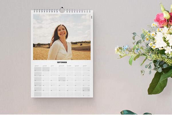 Kalender aan de muur met foto van lachende vrouw in witte blouse op veld onder blauwe lucht en bloemen rechts